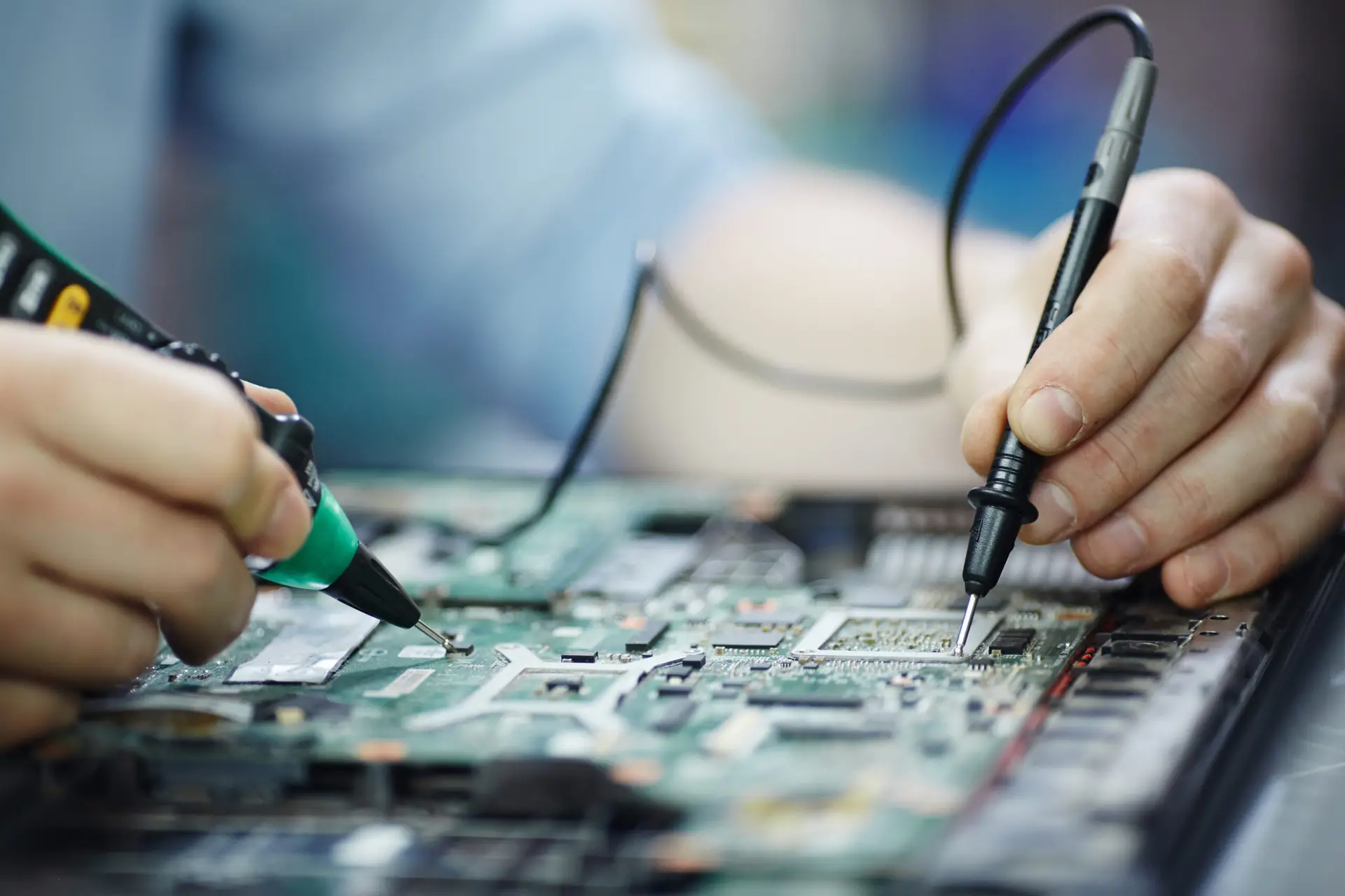 Closeup shot of male hands testing electric current voltage in circuit board of disassembled laptop using multimeter tool on table in maintenance shop